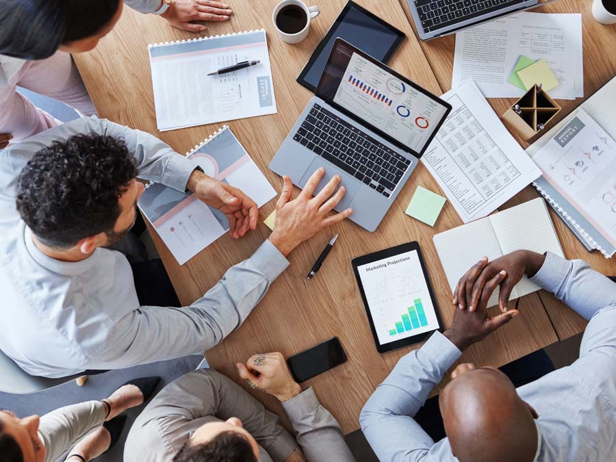 Man blickt von oben auf einen Tisch mit mehreren Unterlagen und einen Laptop. Um den Tisch sitzen einige Personen, die einen Businessplan erstellen wollen.